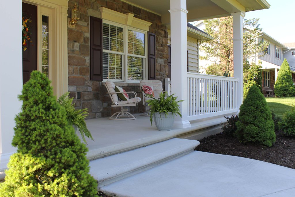 Close up of covered front porch has potted plants, inviting furniture, sidelight on front door